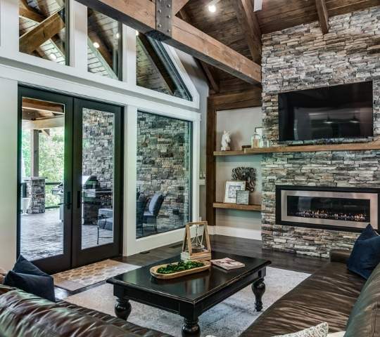 Photo of great room space with exterior wall of window and glass doors opening to extensive outdoor living space with grilling and dining area. The great room has a large gas stone fireplace, exposed wood a-frame beams and exposed wood ceiling.