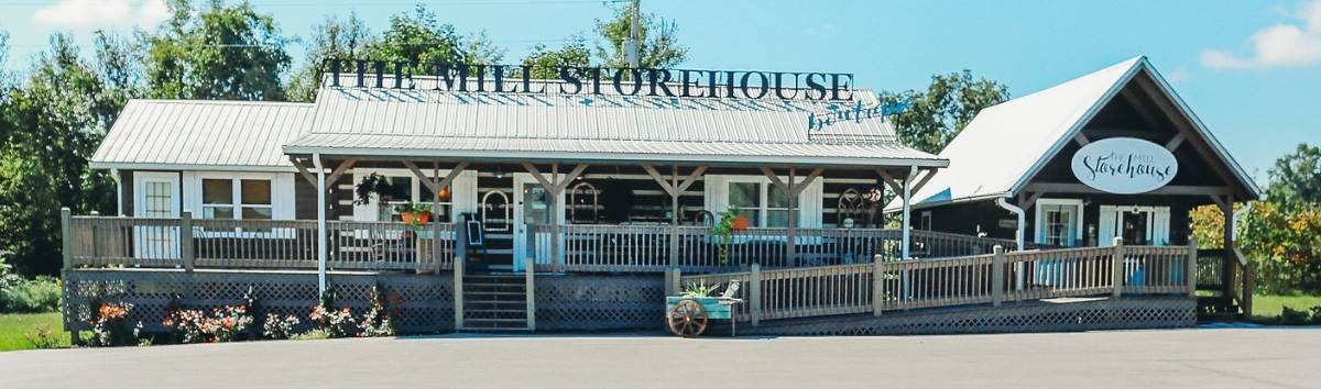 Photo of The Mill Storehouse with a metal roof and chink style dark logs. A long porch extends to a separate smaller building that houses a specialty shop.