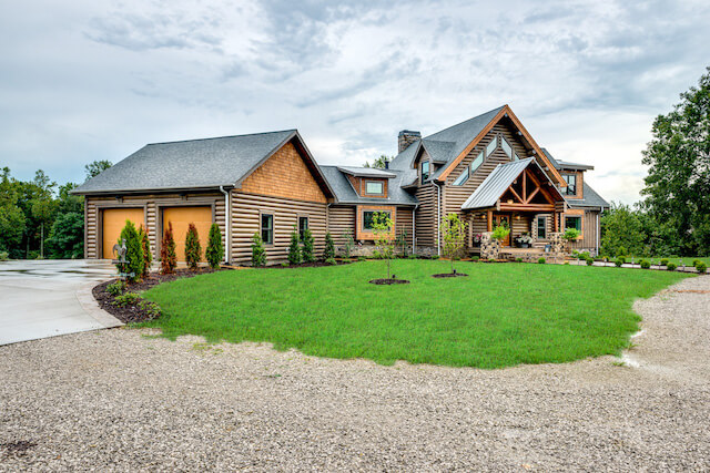 Photo of large log home with double a-frame front entrance covered porch and front porch extending the entire length of the front of the home. Stone front steps and pillars support the porch roof. the home is log chink style.