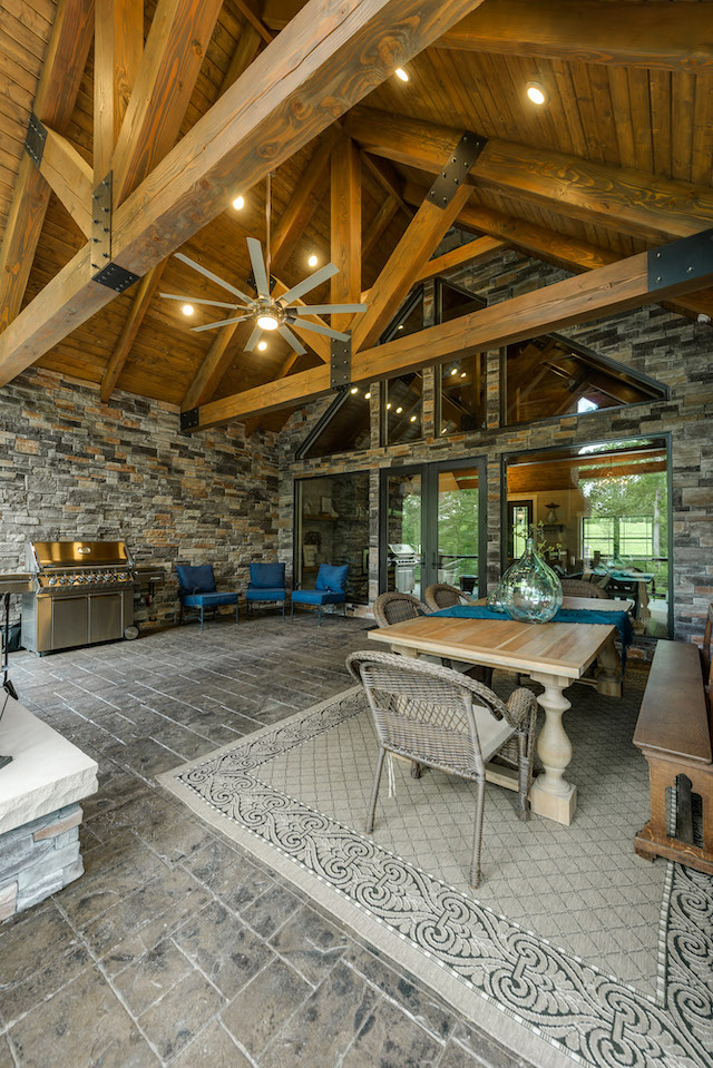photo of porch with a-frame beams and wood ceiling and stone floor. Walls of stone and ceiling fans make the outdoor dining space luxurious